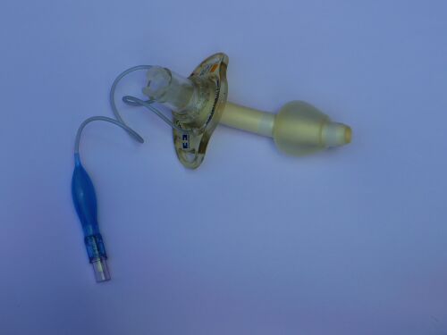 A cuffed tracheostomy tube lying on a pale surface, showing its curved cannula, neck flange, inflated cuff and a blue pilot balloon on a thin inflation line.