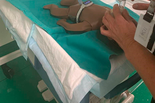 A newborn resuscitation manikin on a draped table being ventilated with a face mask; a heart-rate meter on its chest shows 163.