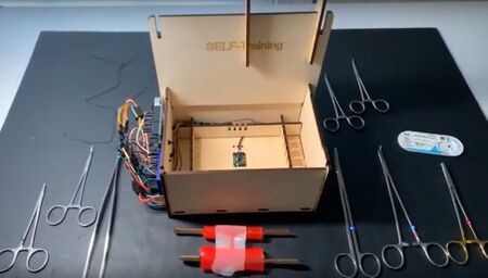 Wooden PDA-ligation training box holding a gelatin-glycerol vascular model, with an electronic sensor display and surgical instruments during a training exercise.