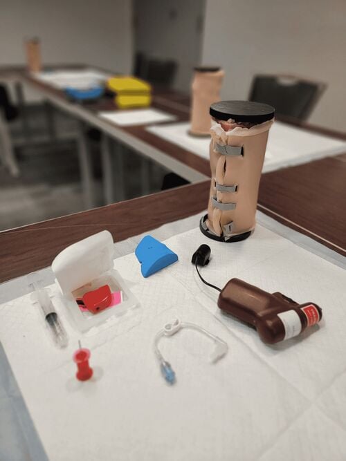 Photograph of the maxSIMIO intraosseous access simulator standing upright on a training table beside an Arrow EZ-IO Power Driver, an EZ-IO Needle Set in its packaging, and a blue landmarking wedge.