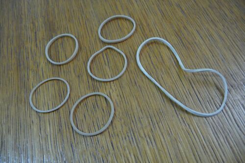 Five pale tan rubber bands of different sizes lying on a wooden desk, four resting as flat loops and one stretched into an elongated lobed shape.
