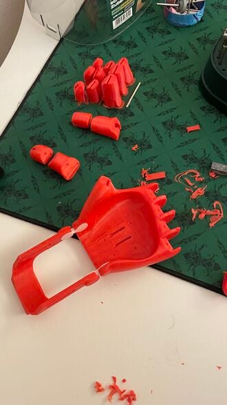 The image shows multiple 3D-printed parts scattered on a table on a cutting mat. The parts are partly cleaned up, but some still have a brim and stringing.
